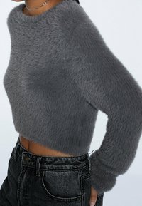 Persona vestida con un suéter corto de pelaje gris oscuro de manga larga y unos jeans de mezclilla negros de cintura alta, de pie contra un fondo liso.