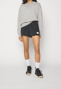 Sudadera gris con un pequeño logo blanco, combinada con pantalones cortos negros que tienen una etiqueta blanca. Calcetines blancos hasta el tobillo y zapatillas deportivas negras con suelas beige.