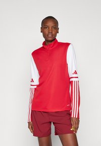 Rød sportstop med en halv-zip krave, hvide �ærmer og tre røde striber på hver ærme. Sammen med mørkerøde shorts med en gul detalje.