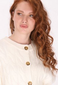 Witte kabelgebreide cardigan met een geribde kraag en drie grote bruine knopen aan de voorkant, met losse golven van rood haar zichtbaar.