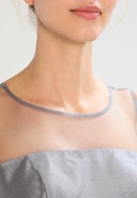 Šedé šaty s průhledným, transparentním výstřihem. Tkanina má jemnou texturu, hladký materiál je pod oblastí hrudníku. Žádné viditelné ozdoby.