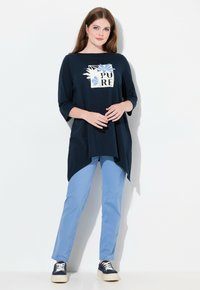 Mörkblå T-shirt med ett blommigt grafiskt mönster som har ordet "PURE." Kombinerad med ljusblå byxor och mörkblå sneakers.