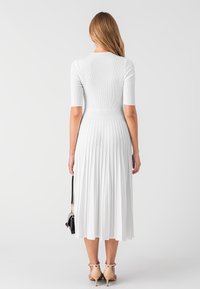 Robe blanche à côtes avec des manches courtes, jupe plissée atteignant le mi-mollet, texture douce et corsage ajusté. Portée avec des talons nude et un sac noir.