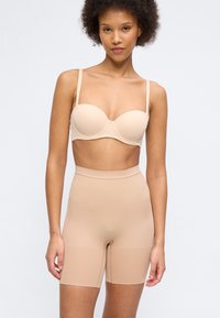 Γυναίκα με σγουρά μαλλιά που φοράει μπεζ στράπλες σουτιέν και σορτς διαμόρφωσης ψηλής μέσης, στεκόμενη μπροστά σε έναν απλό ανοιχτόχρωμο φόντο.