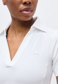 Femme portant un polo blanc à col avec un motif texturé subtil et un petit logo sur le côté gauche de la poitrine.