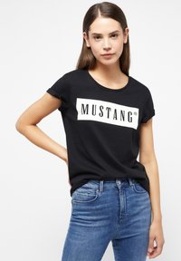 Černé tričko s krátkým rukávem, které má bílý obdélníkový grafický prvek s výrazným nápisem "MUSTANG", v kombinaci s modrými džíny.