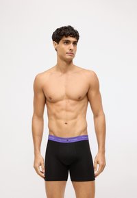 Czarne bokserki z purpurowym pasem, na którym widnieje logo "TOMMY HILFIGER". Materiał wydaje się gładki i dopasowany do ciała.