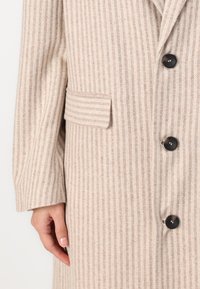 Beige herringbone coat featuring a single black button, a side pocket, and long sleeves with a relaxed fit. Textured fabric finish.
