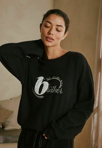 Schwarzer Pullover aus strukturiertem Stoff, mit weitem Ausschnitt und lockerer Passform. Mit prägierten, weißen Buchstaben "6 von Ted Baker." Lange Ärmel und lässige Form.