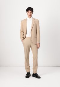 Costume beige à coupe ajustée, comprenant un blazer à bouton unique et un pantalon assorti, associé à une chemise blanche avec col et des chaussures noires.