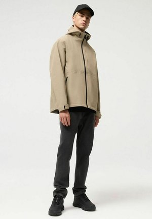 Młody mężczyzna ubrany w beżową kurtkę z kapturem, czarną czapkę, ciemnoszare spodnie i czarne trampki, stojący na tle jasnego, jednolitego tła.