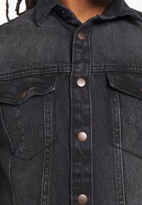 Zwarte denimjas met koperen knopen, met voorste klepzakken en een versleten uitstraling op de stof. Klassieke westerse ontwerpelementen.