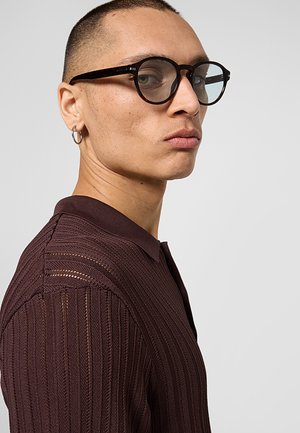 Persona con capelli corti che indossa occhiali rotondi scuri, un orecchino a cerchio d'argento e un maglione marrone scuro strutturato, che guarda di lato.