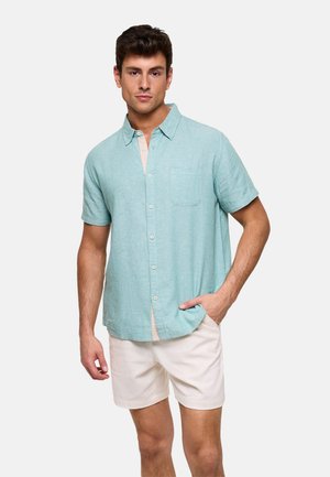 Hombre con camisa de manga corta de botones azul claro sobre camiseta beige y pantalones cortos blancos, de pie con una mano en el bolsillo.