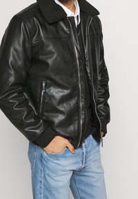 Czarna skórzana kurtka z kołnierzem z owczej skóry, zamkiem błyskawicznym z przodu i kieszeniami, ściągaczami na mankietach oraz gładką fakturą. Noszona na ciemnym swetrze i jasnych dżinsach.