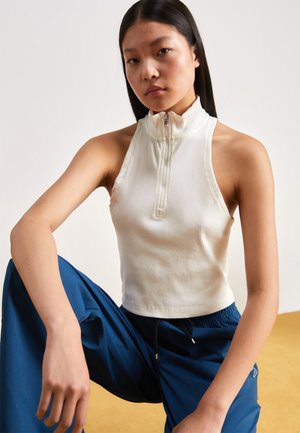 Fiatal nő, hosszú fekete hajjal, ujj nélküli fehér cipzáros felsőt és kék nadrágot viselve, ül egy térdét felhúzva egy sima háttér előtt.