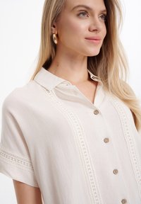 Camicia button-up beige chiaro realizzata in tessuto testurizzato, con colletto e dettagli in pizzo lungo la parte anteriore, dotata di bottoni in legno.