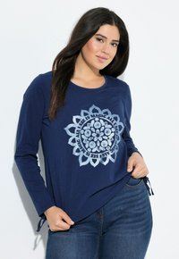 Chemise à manches longues bleu marine avec un design graphique mandala argenté. Tissu doux avec des liens sur les côtés au niveau de l'ourlet pour plus de détail. Portée avec un jean bleu.