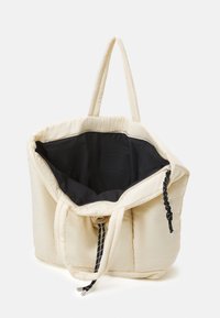Sac fourre-tout beige rembourré avec intérieur noir et fermeture à cordon tressé noir. Dispose de deux poignées robustes pour le transport.