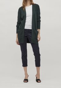 Cardigan verde oscuro con apertura frontal, puños acanalados y dos bolsillos delanteros, combinado con una blusa blanca y pantalones recortados en azul marino. Sandalias negras de tacón.