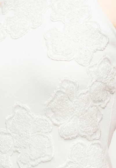 Λευκό ύφασμα με υφασμάτινα κεντήματα από δαντέλα με υφή λουλουδιών και μικρές λεπτομέρειες από χάντρες σε ένα λείο, εφαρμοστό ένδυμα.