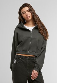 Hoodie cropped in cotone verde scuro con zip frontale, cappuccio attaccato e due tasche frontali. Abbinato a pantaloni in denim neri.