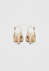 Beige hælede sandaler med et slankt, åbent bagside-design, ankelrem, spids tå og en glat overflade. Tynd hæl med en kontrasterende sort spids.