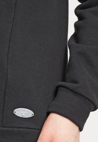 Černá mikina vyrobená z měkké tkaniny, s detailním pohledem na manžetu a stříbrnou visačkou "True Religion" na boku.