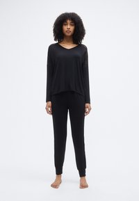 Μαύρο σετ άνετης ενδυμασίας που περιλαμβάνει μπλούζα με μακριά μανίκια, V-λαιμόκοψη και ελαφρώς κομμένη σχεδίαση, σε συνδυασμό με αντίστοιχο παντελόνι με ελαστική μέση.