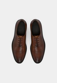 Chaussures en cuir marron à finition lisse, avec un bout arrondi, cinq œillets, et une semelle en caoutchouc. Coutures décoratives le long des côt és.