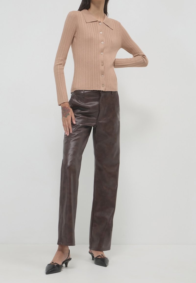 Femme portant un haut beige côtelé à manches longues boutonné, un pantalon en cuir marron foncé et des talons bas noirs pointus sur fond blanc.