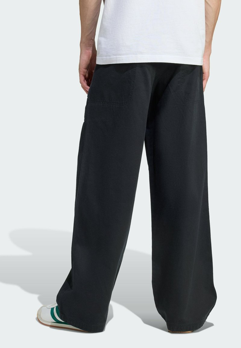 Persoon in losse zwarte broek, wit shirt en witte sneakers met groene strepen, staand voor een effen lichte achtergrond.
