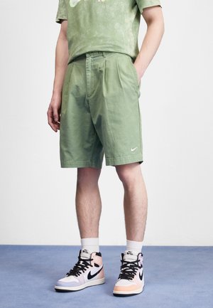 Shorts en coton verts avec des plis, des poches latérales et un logo Nike. Associés à des baskets montantes Nike pastel et à un t-shirt assorti en tie-dye vert.