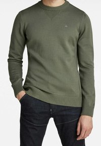 Olivgrüner Pullover mit geripptem Muster, Rundhalsausschnitt und kontrastierenden Strickmustern an Schultern und Saum. Kombiniert mit dunkler Jeans.