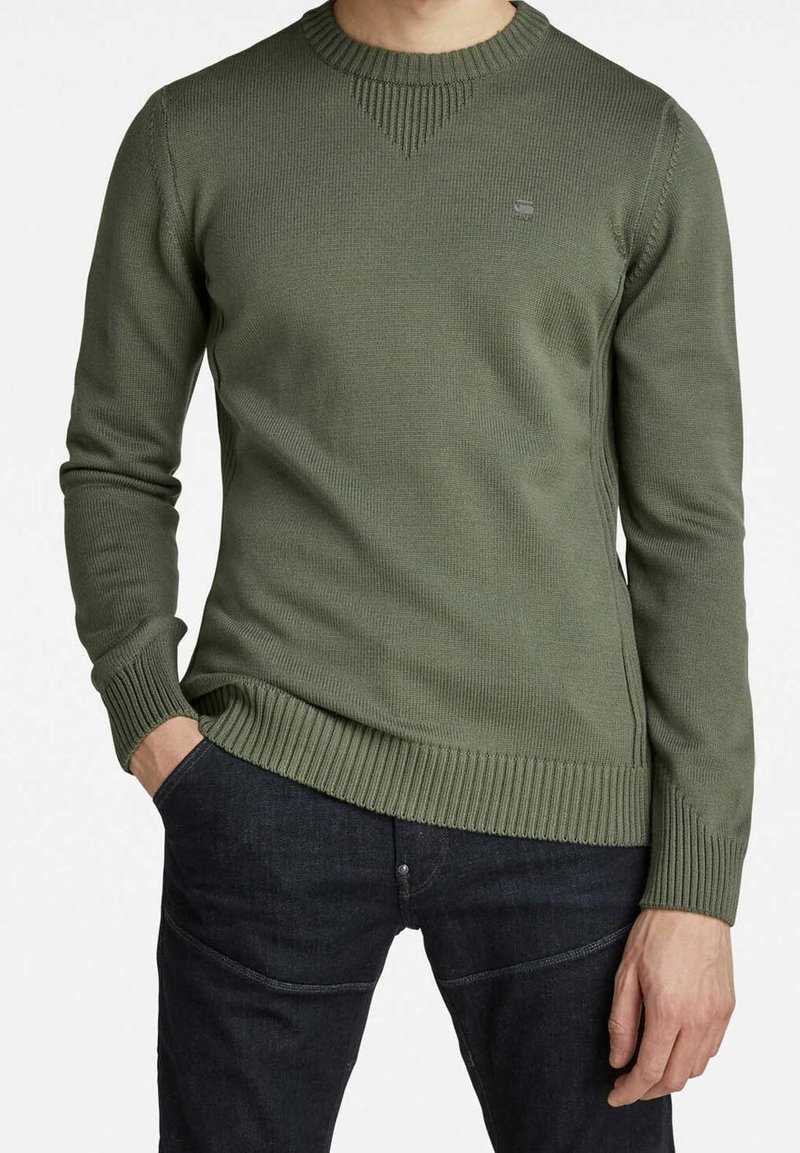 Olivgrüner Pullover mit geripptem Muster, Rundhalsausschnitt und kontrastierenden Strickmustern an Schultern und Saum. Kombiniert mit dunkler Jeans.