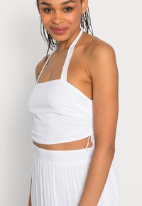 Top cropped branco com alças, em tecido texturizado, com laços ajustáveis nas laterais e um corpete justo, combinado com uma saia esvoaçante branca.