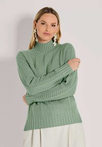 Frau in grünem strukturiertem Rollkragenpullover und cremfarbenem Rock, accessorized mit großen, blumigen weißen Ohrringen, posiert mit verschränkten Armen.