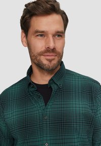 Groene geruite button-up shirt met een subtiel ruitpatroon, zwarte knopen en een klassieke kraag. Gedragen over een zwart T-shirt.