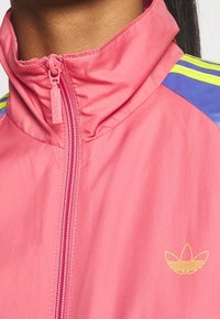 Detailní záběr osoby v růžové bundě Adidas s límcem na zip, žlutým logem Adidas a modrými a žlutými pruhy na ramenou.