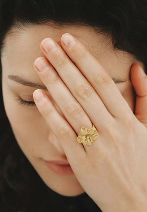 Mujer con los ojos cerrados cubriéndose la cara con una mano, que lleva un anillo dorado en forma de flor con una pequeña piedra preciosa en el dedo medio.