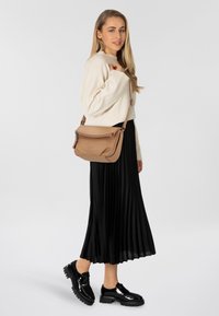 Braune Leder-Crossbody-Tasche mit Klappe und Reißverschluss. Das Model trägt einen cremefarbenen Pullover und einen schwarzen Plisseerock sowie glänzende schwarze Schuhe.