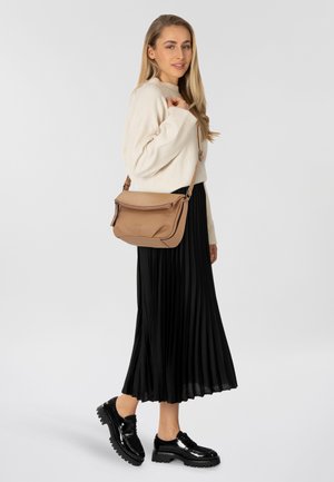Braune Leder-Crossbody-Tasche mit Klappe und Reißverschluss. Das Model trägt einen cremefarbenen Pullover und einen schwarzen Plisseerock sowie glänzende schwarze Schuhe.