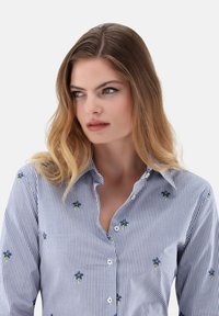 Striped button-up shirt in light blue and white, featuring small blue floral embroidery and a classic collar. Smooth fabric texture.