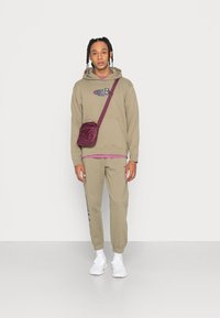 Khaki kapuce ar logo, saskaņota ar atbilstošām jogger biksēm. Baltas sporta kedas un mazs, bordo krāsas crossbody soma. Vienkāršs, ikdienišķs dizains.