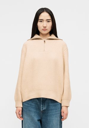 Γυναίκα με μακριά σκούρα μαλλιά που φοράει μπεζ πλεκτό πουλόβερ με φερμουάρ έως το στήθος και γιακά, συνδυασμένο με χαλαρό μπλε τζιν, σε λευκό φόντο.