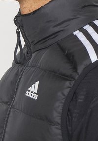 Detailní záběr černé zateplené vesty Adidas s vysokým límcem a částečným zipem, s bílými třemi pruhy na rameni a logem na hrudi.