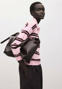 Frau in einem pink- und burgunderfarbenen gestreiften Langarmshirt, die zwei große schwarze Ledertaschen über der Schulter trägt und vor einer weißen Wand steht.