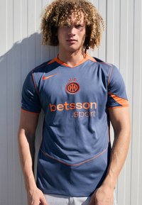 Maillot de sport bleu marine avec des accents orange. Il comprend un col en V, des manches courtes et des logos pour Betsson et un emblème de l'équipe.