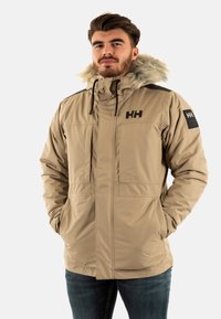 Giacca isolante beige con cappuccio foderato in pelliccia, tasche frontali e dettagli neri sulle spalle. Presenta un logo sul petto e sulla manica.