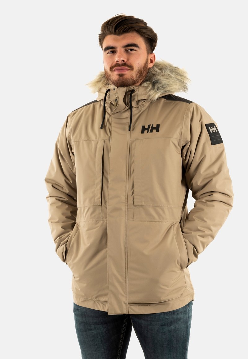 Giacca isolante beige con cappuccio foderato in pelliccia, tasche frontali e dettagli neri sulle spalle. Presenta un logo sul petto e sulla manica.